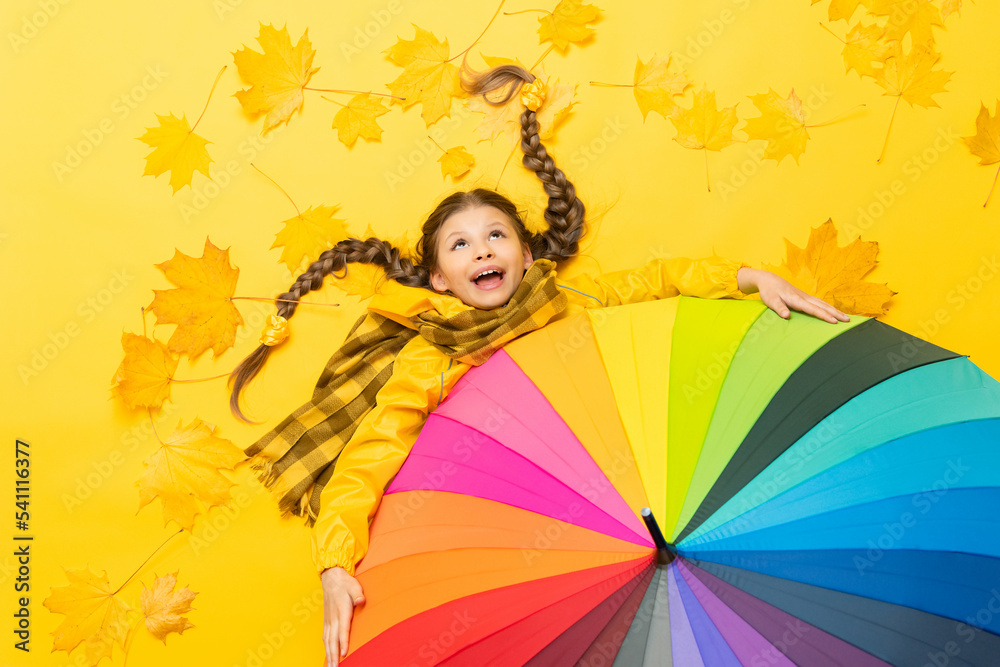 A little girl in a raincoat with a scarf lies on autumn, yellow, maple leaves and holds onto a colorful colorful umbrella. A funny child with pigtails is enjoying the autumn season. A little girl in a raincoat with a scarf lies on autumn, yellow, maple leaves and holds onto a colorful colorful umbrella. A funny child with pigtails is enjoying the autumn season.