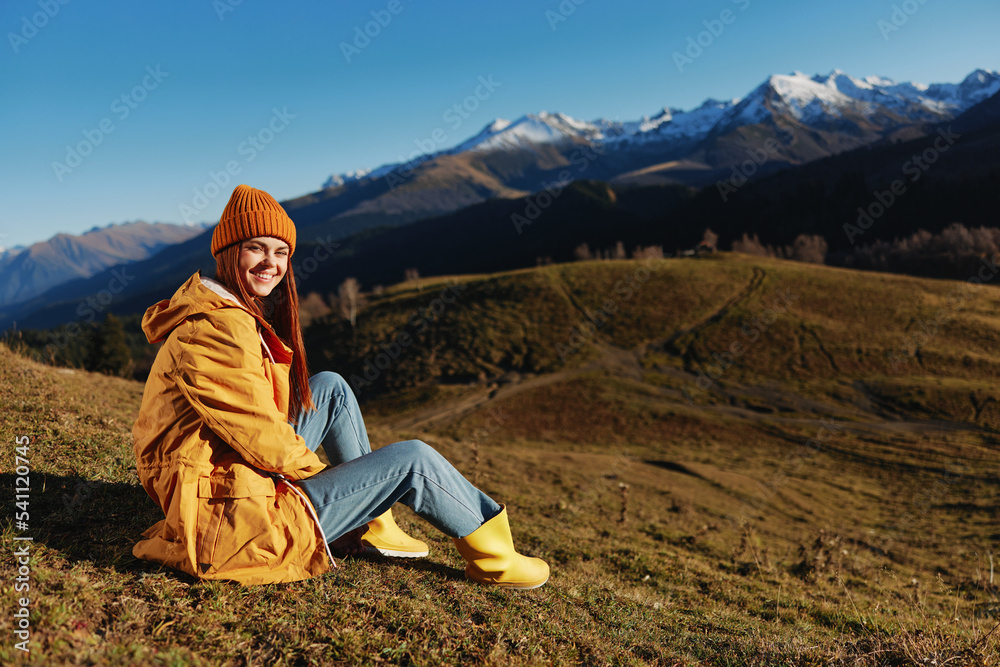 Woman sitting on a hill on the grass rest smile with teeth looking at the mountains in the snow in the winter in a yellow raincoat and jeans happy sunset trip on a hike, freedom lifestyle 