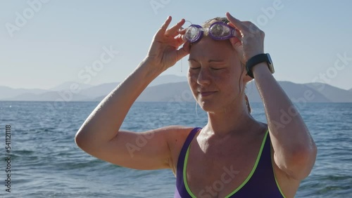 young woman on coast of sea or ocean takes off her purple diving goggles that match her swimsuit coloe and look at camera. girl has finished bathing and takes off her accessories. photo