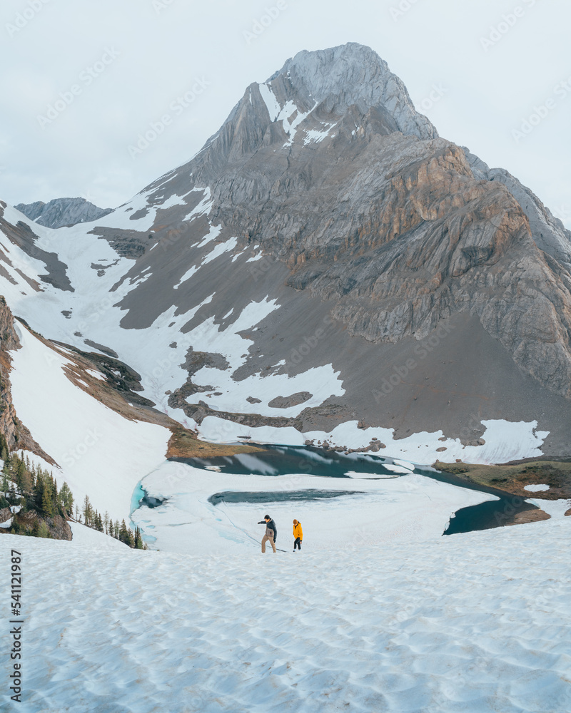 two hikers hiking a snowy mountain with a beautiful lake and mountain on the background