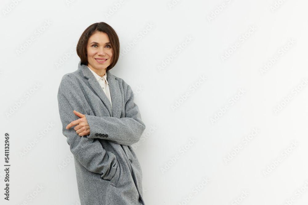 a horizontal portrait on a light background in a long gray coat stands smiling at the camera in a closed pose a horizontal portrait on a light background in a long gray coat stands smiling at the camera in a closed pose