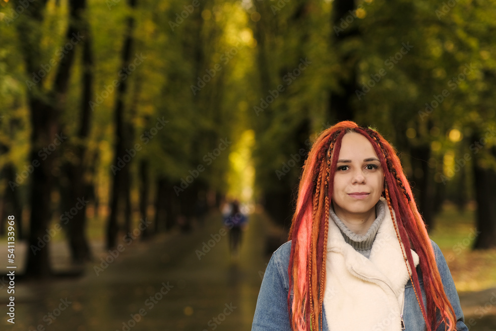 Portrait of young woman with ginger dreadlocks posing in forest park. Pretty female with bright hairstyle in warm denim jacket on walk.