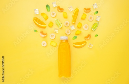Bottle of orange fruit juice under cutted different fruits and ice in yellow background