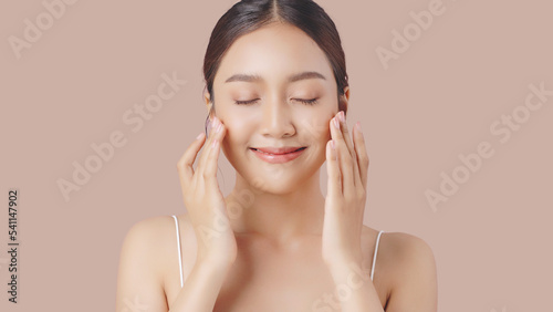 Face cream commercial advertising concept. Young beautiful Asian woman massages her face gently with fingertips isolated over beige brown background.