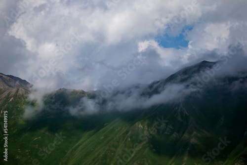 Dark atmospheric surreal landscape with a dark rocky mountain peak in low clouds in a gray cloudy sky. A gray low cloud on a high peak. High black rock in low clouds. Surreal gloomy mountains.