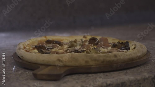 Professional chef adds grated parmesan and cuts traditional Capricciosa with pizza knife on wooden board at a marble countertop. Pizzaiolo cooks a traditional Italian dinner in a modern restaurant kit