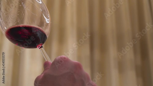 Wine degustation. Man holds a glass with a long leg, swirls a wineglass and inhales the aroma of red wine. Noble drink rotates and covers the walls of transparent goblet in a warm interior.