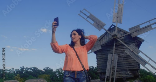 Wallpaper Mural A young attractive brunette woman wearing jeans and a blouse takes a selfie on a smartphone in front of an old wooden windmill. Torontodigital.ca