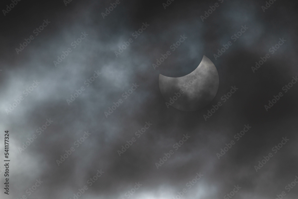 Zaćmienie słońca. Zachmurzone niebo, zza cienkiej warstwy chmur widać słońce w trakcie częściowego zaćmienia. Górna część słońca zasłonięta jest przez księżyc. Na słońcu widoczne są plamy.