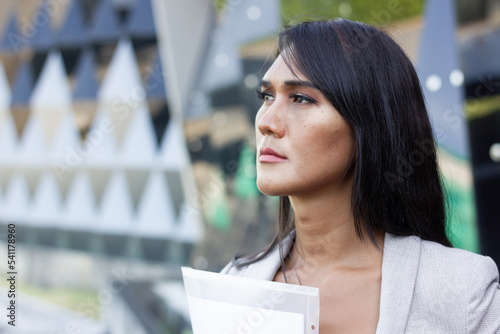 Dark hair Thai businesswoman with determination look holding folder in urban city. Serious Asian transgender person portrait with files in hand, thinking about project ideas by office building