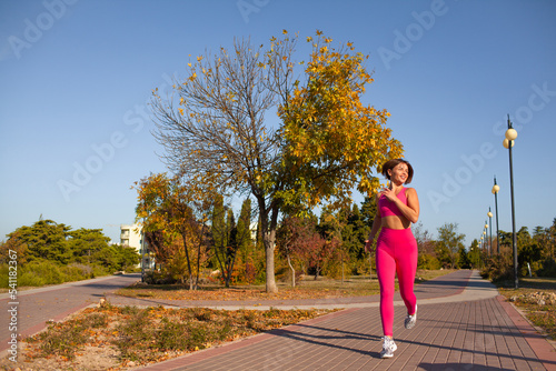 A beautiful woman leads a healthy lifestyle, practises sport in nature. The girl is wearing a pink fuchia tracksuit and running in the parkon the street against the background of trees photo