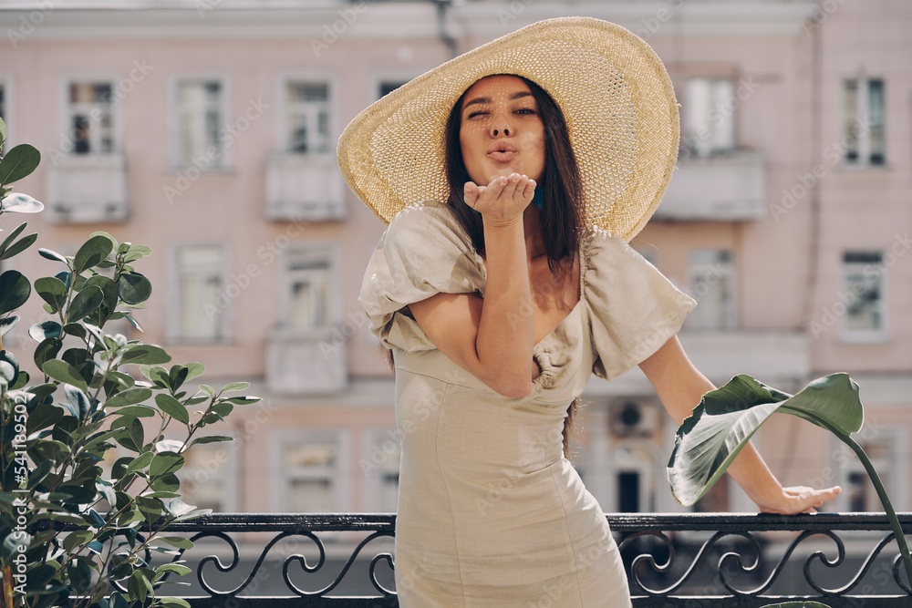 Full length of beautiful young woman in elegant hat blowing a kiss while relaxing on the balcony