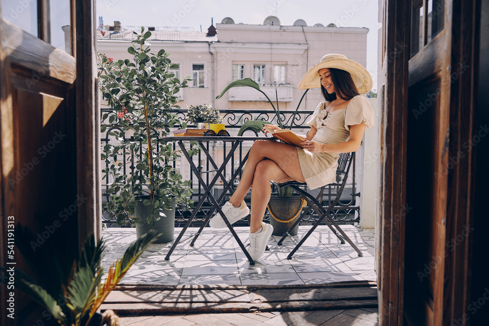 Happy young woman in elegant hat reading book while relaxing on the balcony