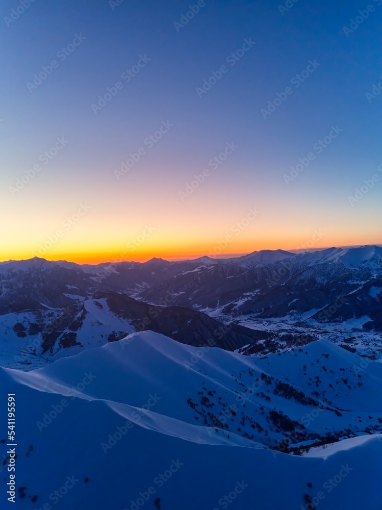 Vertical aerial panorama of snowy mountain ridge on winter sunrise. Stunning mountains range covered with snow powder on ski resort at sunset. Caucasus mountain peaks skyline in the dusk golden hour.