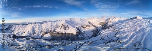 Wide aerial panorama of snowy mountain ridge on winter sunrise. Stunning mountains range covered with snow powder on ski resort at sunset. Caucasus mountain peaks skyline in Georgia - Gudauri.