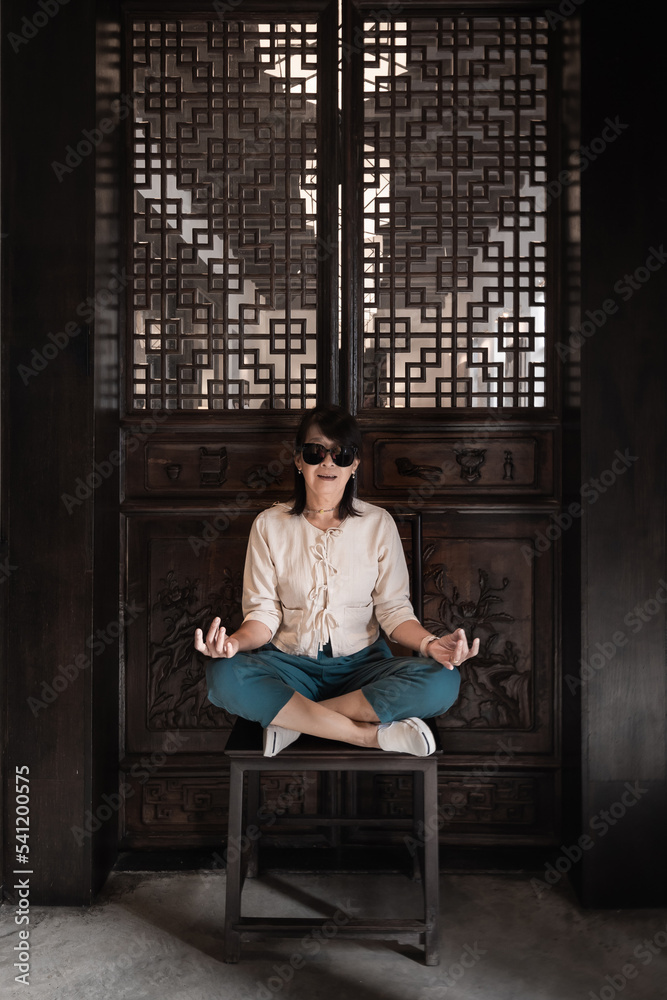 Beautiful portrait of an Asian senior woman sitting on an old wooden chair in a casual clothing fashion dress in a retro classic house background vintage style.