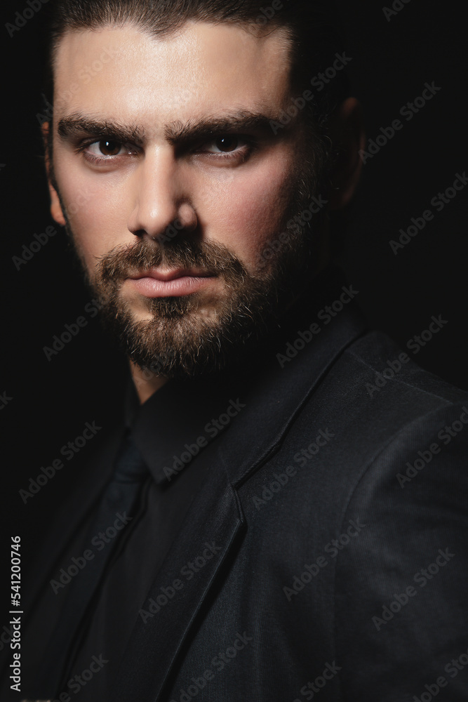 Fototapeta premium Portrait of a white Caucasian male brunet in a black classic suit, black shirt and black tie over a black background. Fashionable portrait of an attractive man with dark hair