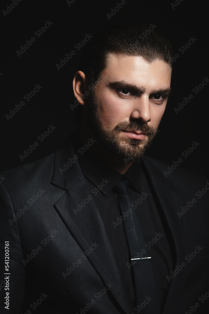 Portrait of a white Caucasian male brunet in a black classic suit, black shirt and black tie over a black background. Fashionable portrait of an attractive man with dark hair