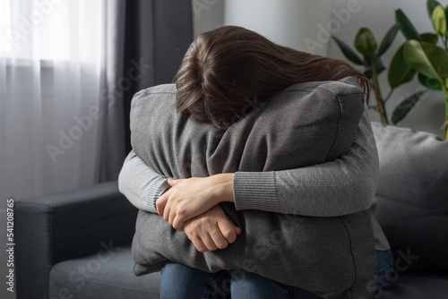 Depressed single young woman holding pillow going through emotional crisis after abuse abortion sitting on couch at alone home. Lonely stressed female in sofa hide from problems of hard cruel world