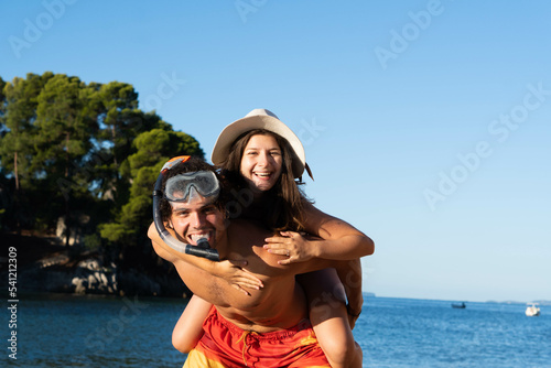 Happy young couple having fun on beach. Boyfriend with snorkeling mask giving piggy back ride to his girlfriend with hat, laughing and enjoying summer at sea.