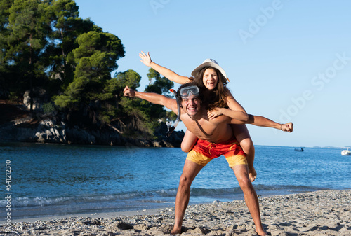 Happy young couple having fun on beach. Boyfriend with snorkeling mask giving piggy back ride to his girlfriend with hat, laughing and enjoying summer at sea.