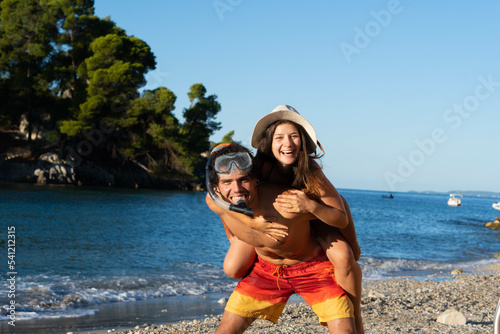 Happy young couple having fun on beach. Boyfriend with snorkeling mask giving piggy back ride to his girlfriend with hat, laughing and enjoying summer at sea.