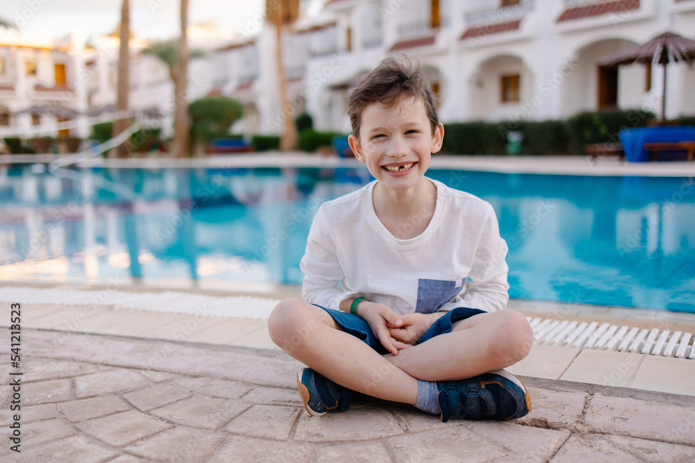 A cheerful child in a long-sleeved T-shirt sits by the pool. Rest in the hotel. Happy holidays. Winter or autumn at the resort. Warm evening. swimming pool is heated. Free childhood. child lost a toot