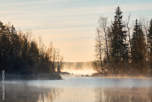 Panoramic view of the forest lake at sunrise. Soft golden sunlight, clear blue sky, reflections on water, fog, frost. Mighty trees. Finland. Nature, seasons, ecology, environment, eco tourism themes © Aastels