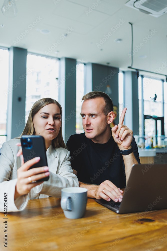 Two male and female colleagues discussing business on laptop, sitting at table in coworking space and talking, young male and female students studying together