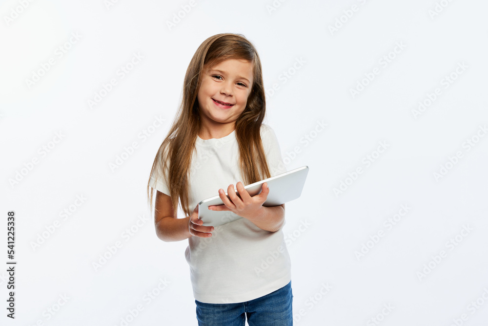 Joyful beautiful little girl, pupil in casual style clothes using tablet isolated over white background. Concept of studying, fashion, back to school Joyful beautiful little girl, pupil in casual style clothes using tablet isolated over white background. Concept of studying, fashion, back to school