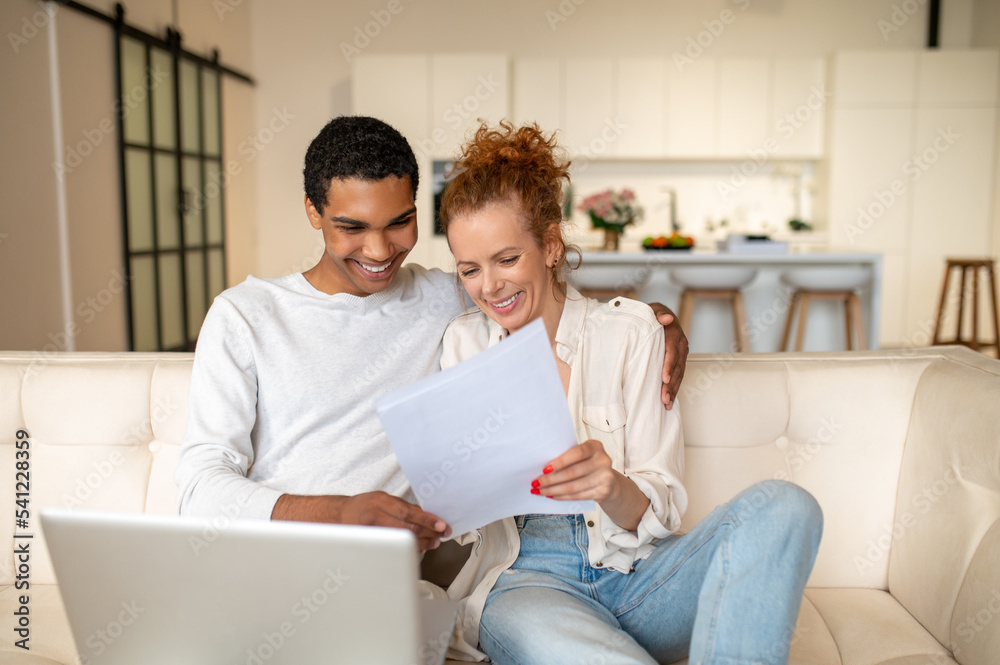 Young man with a laptop sitting on the sofa, his wife standing next to him Young man with a laptop sitting on the sofa, his wife standing next to him