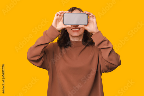 Portrait of young woman holding smartphone over eyes and smiling over yellow background