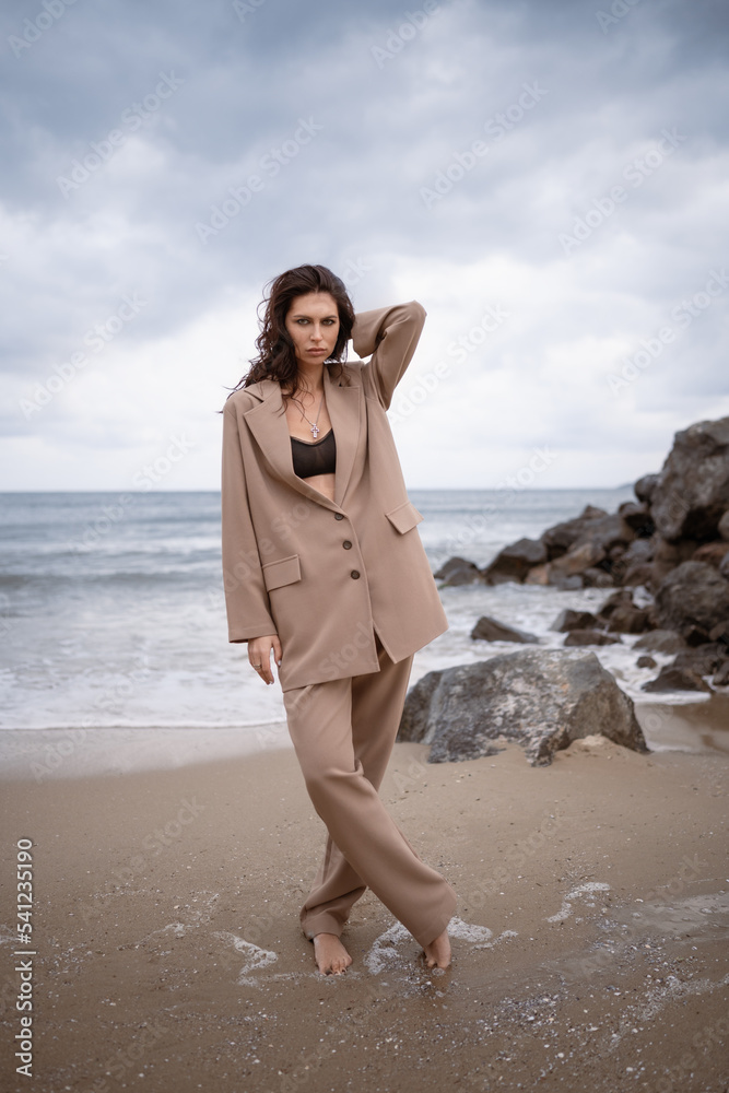 Trendy casual spring  autumn summer outfit. Street fashion. Girl on the nature on dark sky background. Fashion photo.Brunette, beige suit. Elegant woman walking. Sand beach. Sea view.