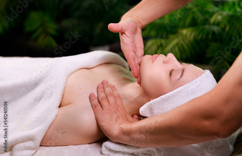 Close-up of a beautiful woman smiling with her eyes closed, receiving a soothing head massage. Young attractive woman enjoying a facial massage at the spa. Skin care, health, body care concept. High
