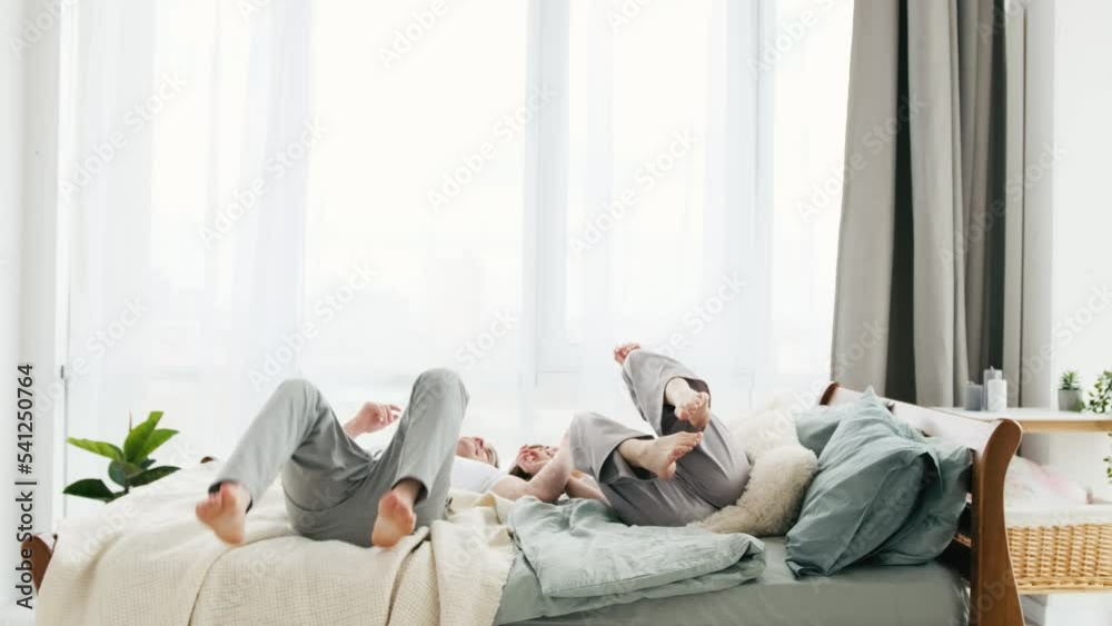 Handsome man and woman wearing pajamas have fun at home. Beautiful couple run in the bedroom and fall down in the bed smiling. Young wife and husband in the morning time in sunny room enjoying moment.