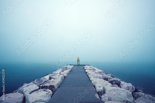 Eine Person mit Knalliger Kleidung steht am Ende Eines Weges der in das Meer ragt und von Wasser umgeben ist. Dichter See Nebel herrscht über dem Meer und macht eine Aussicht unmöglich.