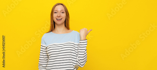 Stylish young smiling woman with fair-haired pointing thumb up to the side, showing advertisement, looking with a happy smile on face, standing over yellow background