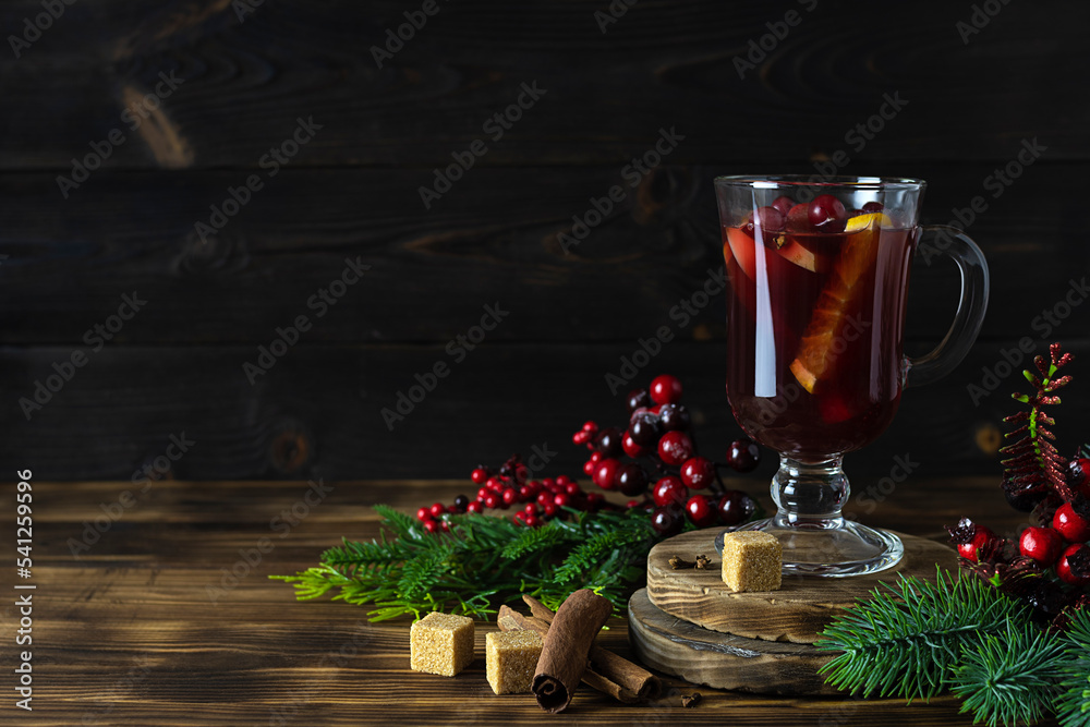 Transparent glass with red fragrant mulled wine on a wooden stand. Red berries, spruce branches and spices in the background. Dark wooden background, space for text