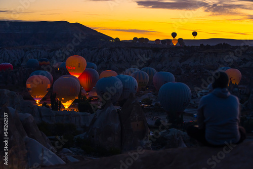 Tourist sitting on top of hill watching glowing hot air ballons flying over Cappadocia at night. Bright orange yellow sky. Horizontal shot. High quality photo