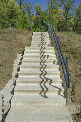 Stone stairs in the park with iron railings and shadow. Landscaping of the park area with trees against the blue sky. Park landscape.