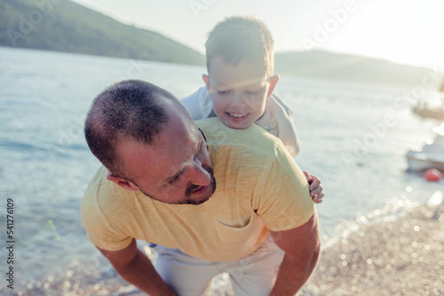 Photo of happy father piggybacking son at sea shore. Man is playing with boy at beach during summer. They are enjoying vacation.....