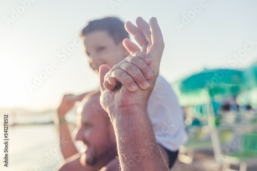 Cute little caucasian boy from behind cheering with arms outstretched while sitting on his father s shoulders at the beach.