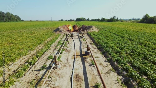 Drone footage of green field with hoses, gas installation. Fire burning in pit. Metal supports, concrete foundation. Daytime. Blue sky, agricultural crops. Industry, business. Production of gas