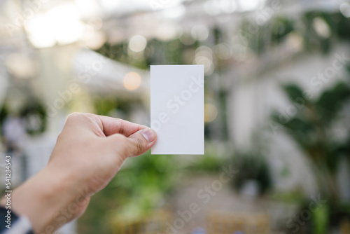 Hand holding a white card with the garden dinner blurry background © JCM