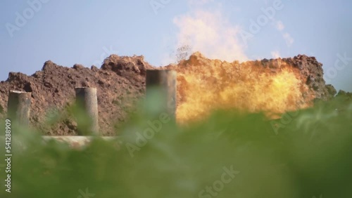 Blurred view of steel tubes and fire. Green grass and blue sky. Tools, equipment for production of gas. Enterprise out of town. Countryside. Rural landscape. Industrial concept