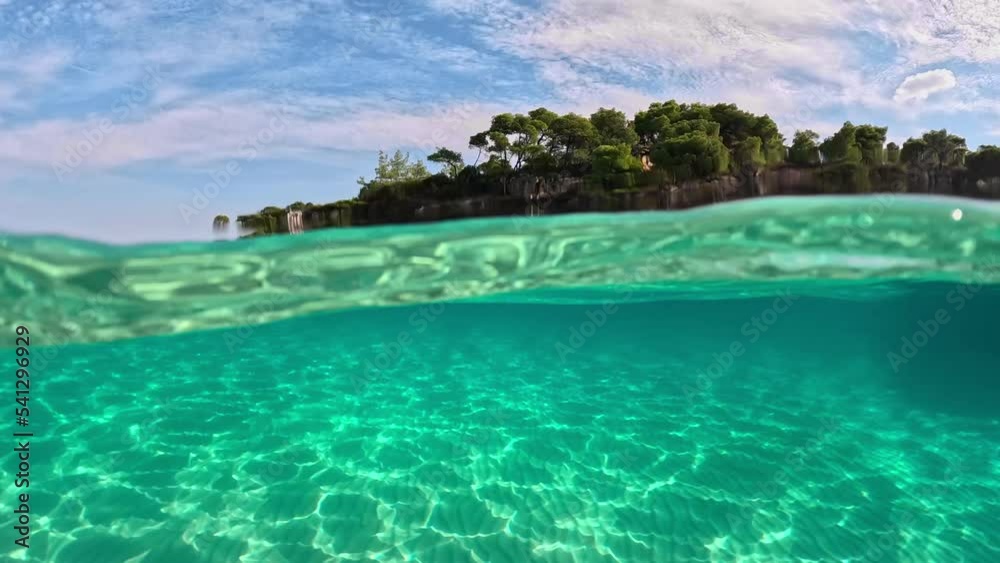 Half underwater slow-motion view of sandy beach bottom and rock with trees. Crystal clear sea water in Sitonia, Halkidiki, Greece. Shot from border of air and water with dome Half underwater slow-motion view of sandy beach bottom and rock with trees. Crystal clear sea water in Sitonia, Halkidiki, Greece. Shot from border of air and water with dome