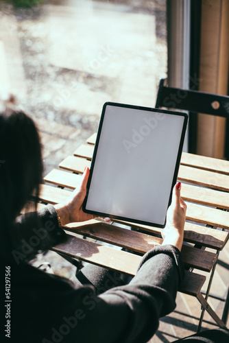 Woman with red nails holding an iPad in a bright room on a wooden table near the window. Work from home