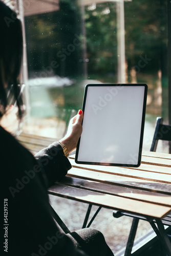 Woman with red nails holding an iPad in a bright room on a wooden table near the window. Work from home