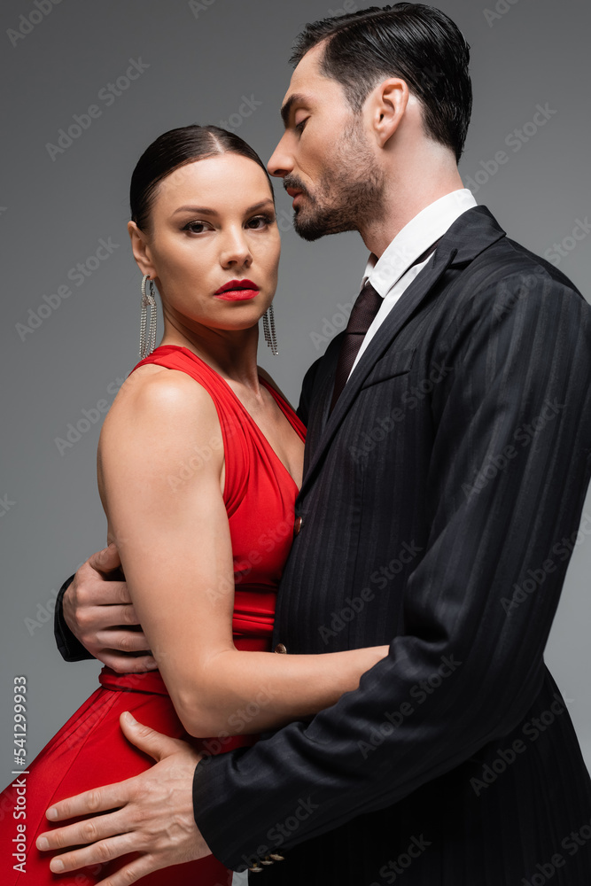 Portrait of woman in red dress looking at camera while hugging elegant boyfriend isolated on grey Portrait of woman in red dress looking at camera while hugging elegant boyfriend isolated on grey