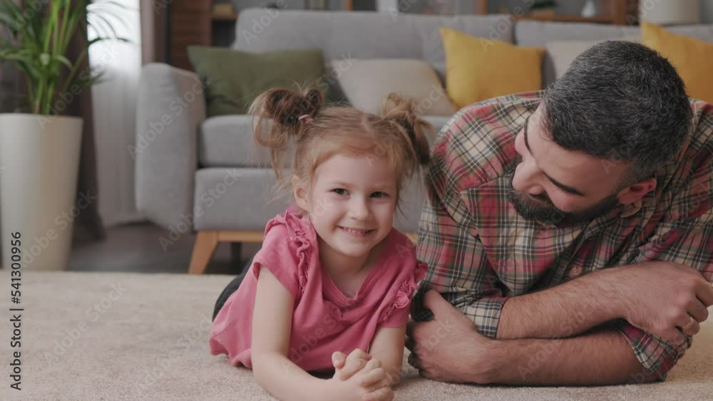 Family portrait at home. Young handsome father with cute daughter lying down on a carpet near the couch. Daddy kissing his liitle girl on the cheek and smiling. Father's day. Family portrait at home. Young handsome father with cute daughter lying down on a carpet near the couch. Daddy kissing his liitle girl on the cheek and smiling. Father's day.